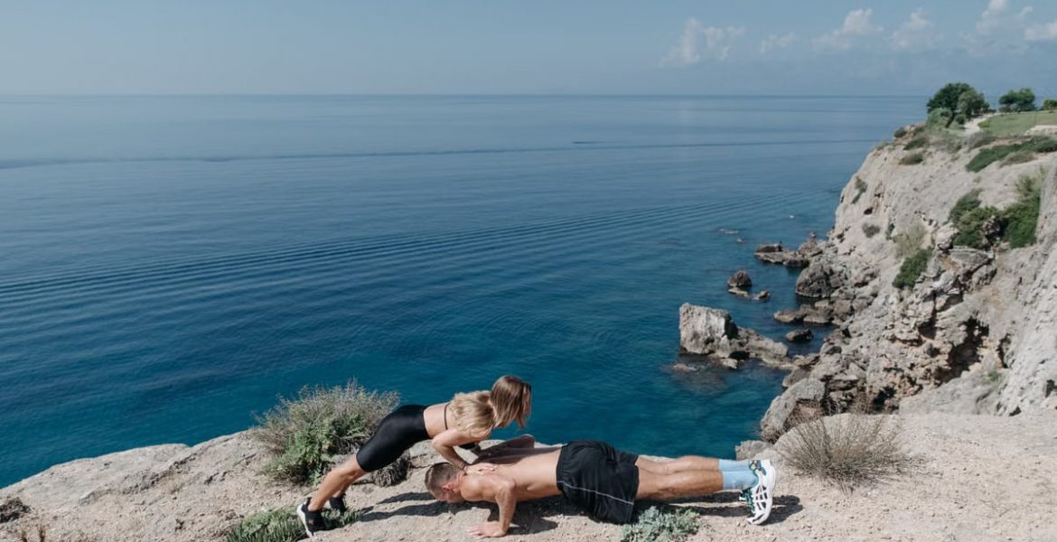 A couple performs a partner workout on a scenic cliffside, overlooking the ocean.