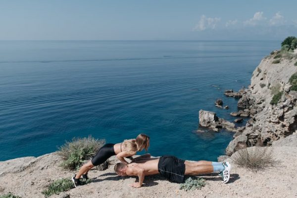 A couple performs a partner workout on a scenic cliffside, overlooking the ocean.
